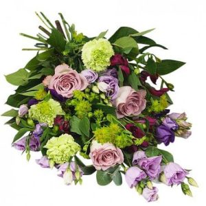 Liggande begravningsbukett med blandade blommor i lila, ljust lila och lime mot gröna blad. Här: rosor, prärierosor, krysantemum (blomsorter kan variera lite i olika butiker, men aktuell blomsteraffär för ditt blombud kommer att göra allt för att efterlikna buketten på bilden så mycket som är möjligt).