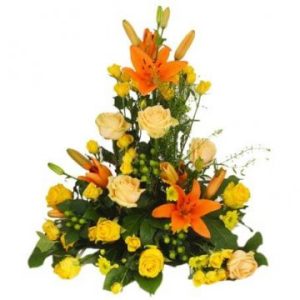 Hög begravningsdekoration med snittblommor i orange och gult på en bas av gröna blad. Här: orange liljor och rosor i olika gula nyanser (blomsorter kan variera lite i olika blomsterbutiker)