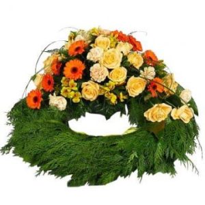 Rund, klassisk begravningskrans i grönt, med blandade blommor i orange, gult och vitt på översta halvan av kransen.